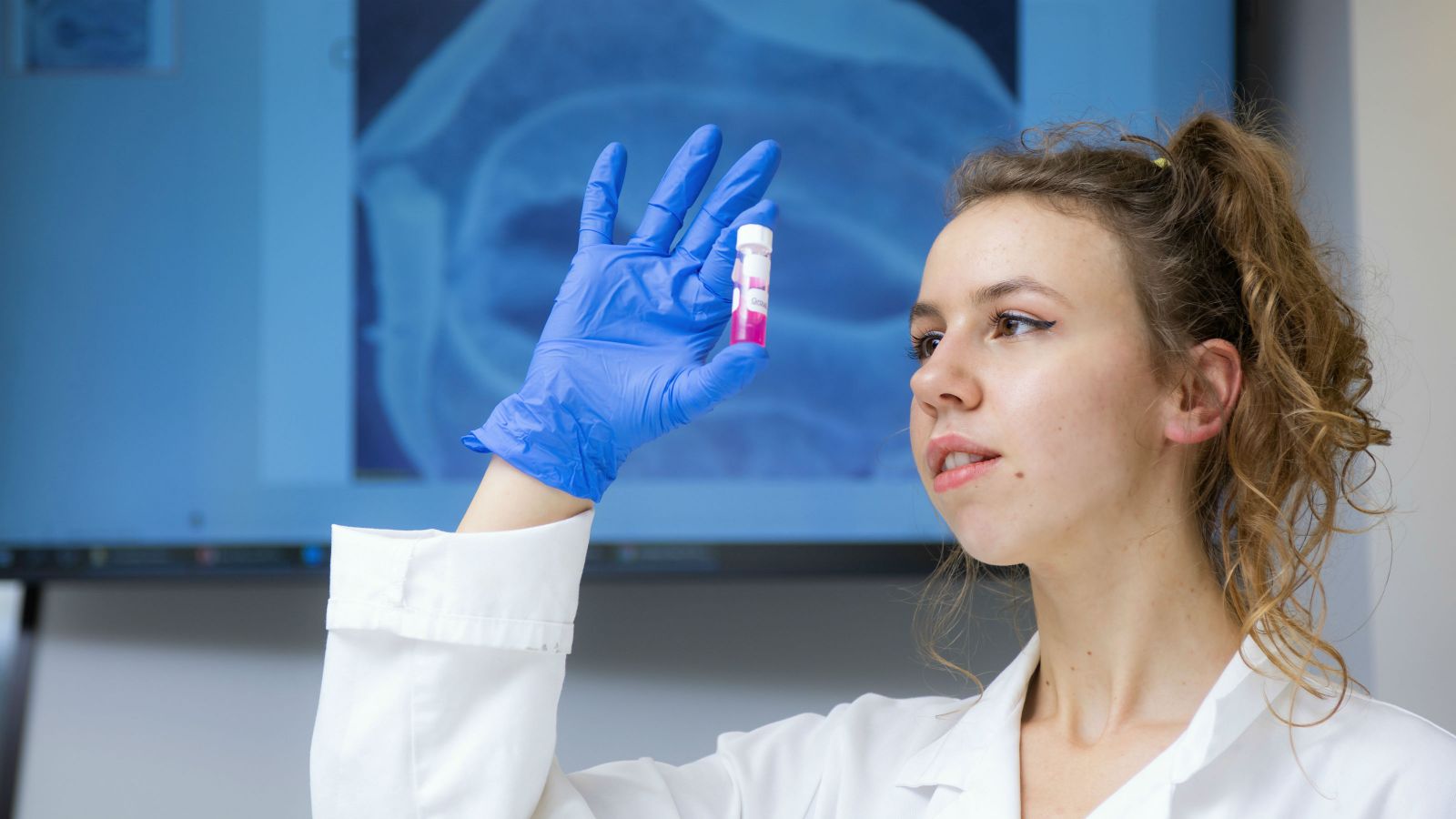 Alinor, wearing a lab coat and a blue plastic glove, holds up a vial of pink liquid. 