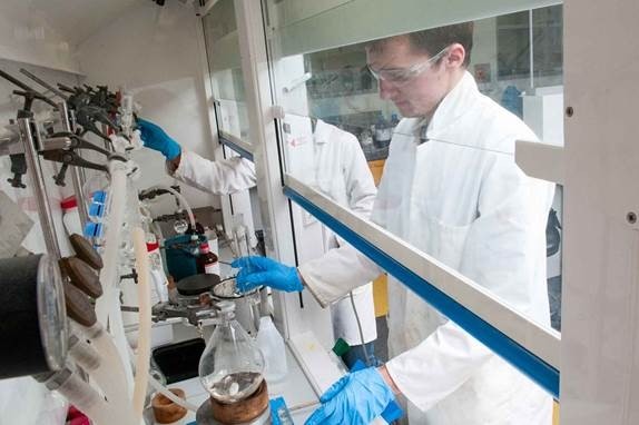 student standing in lab gear conducting experiment using equipment inside fumehood