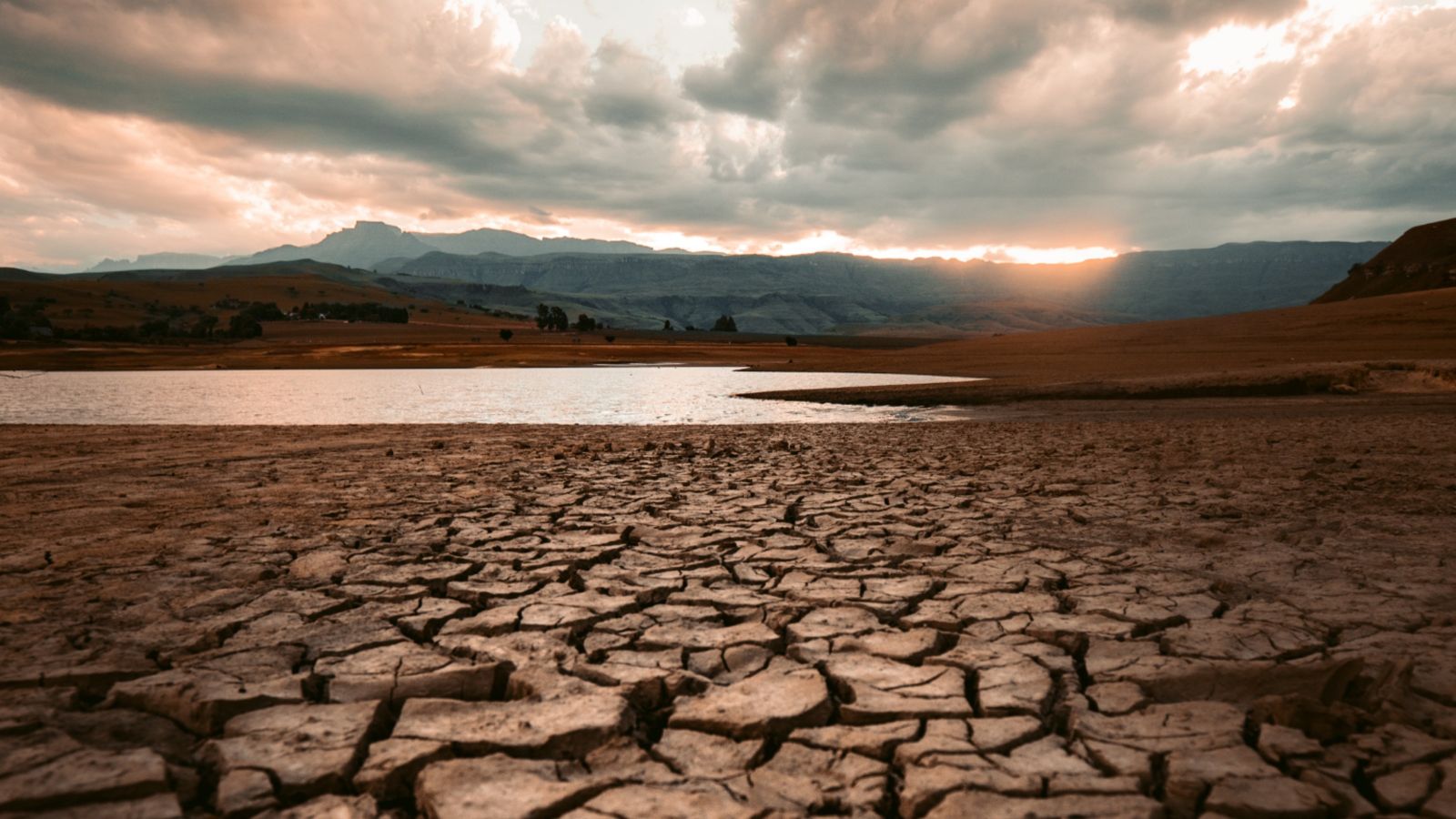Dry cracked lake bed with little water against a background of a setting sun over mountains.