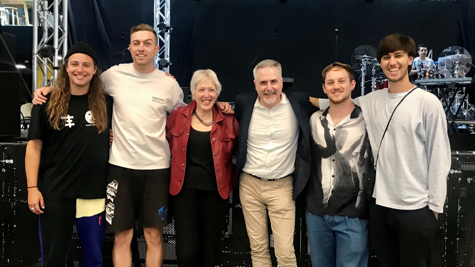 5 men and 1 women stand arms around shoulders in front of a stage with a drummer in the background. Left to right: Ben O'Leary, Matt Beachen, Sally Jane Norman, Lance Philip, Sam Thomson, Shaan Singh.