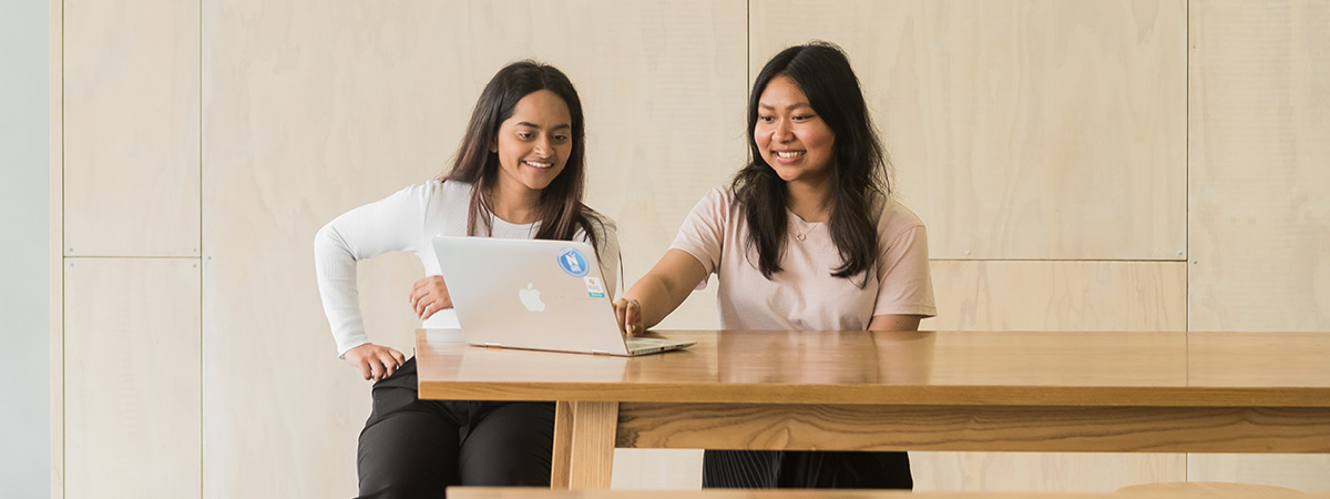 Two female students seated at a wooden table using a laptop. banner image