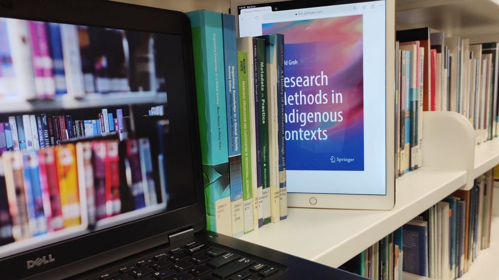 Multicoloured spines of selection of print books on a library shelf with a black laptop displaying eBooks, and a white tablet, displaying the front cover of an eBook, emerging from between the print books.