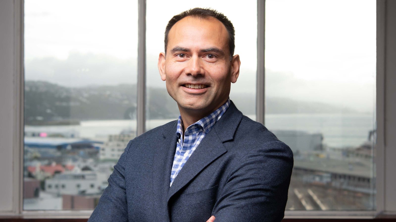 An Executive MBA student poses for a photo with his arms crossed in front of a window overlooking Wellington Harbour.