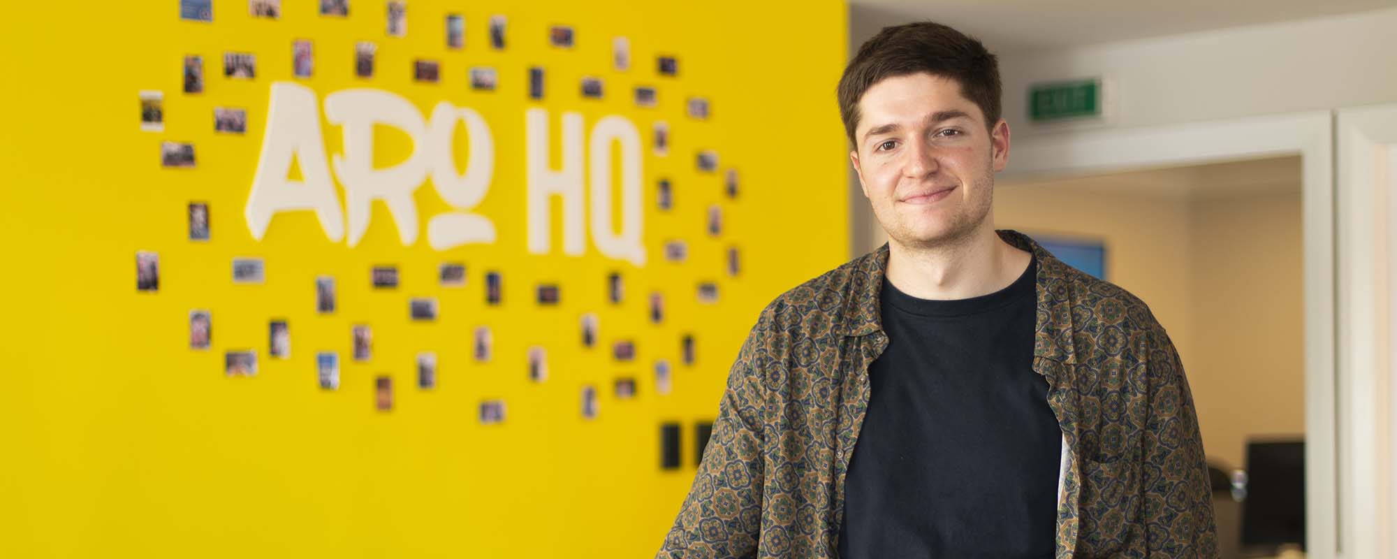 Daniel Raats stands smiling in front of a wall with 'Aro Digital' written in white on a yellow background.