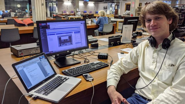 Man sits facing camera with a laptop and desktop computer behind him with the image of a cassette tape on the screens.