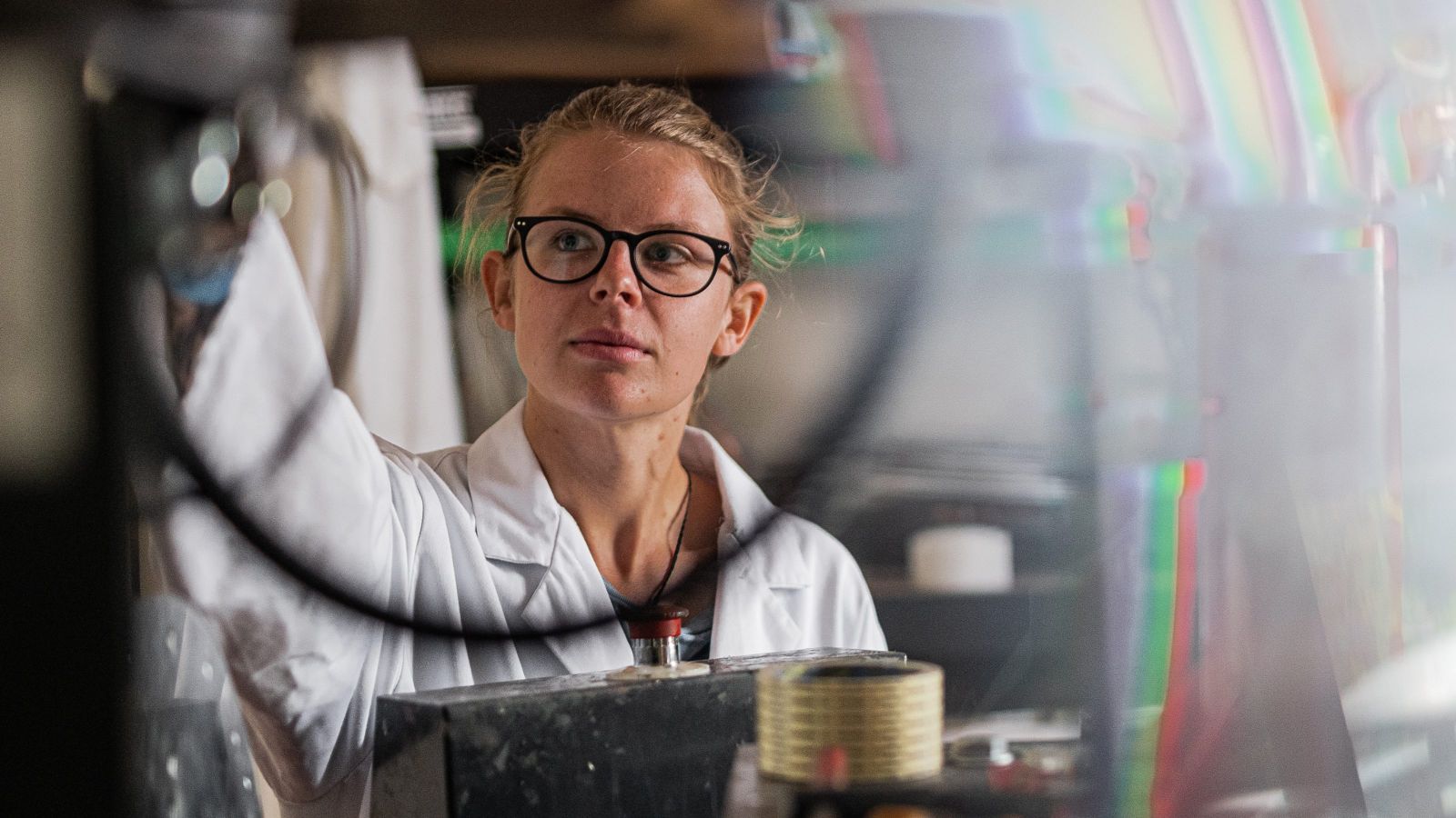A female scientist wearing a white lab coat viewed through wires and machinery.
