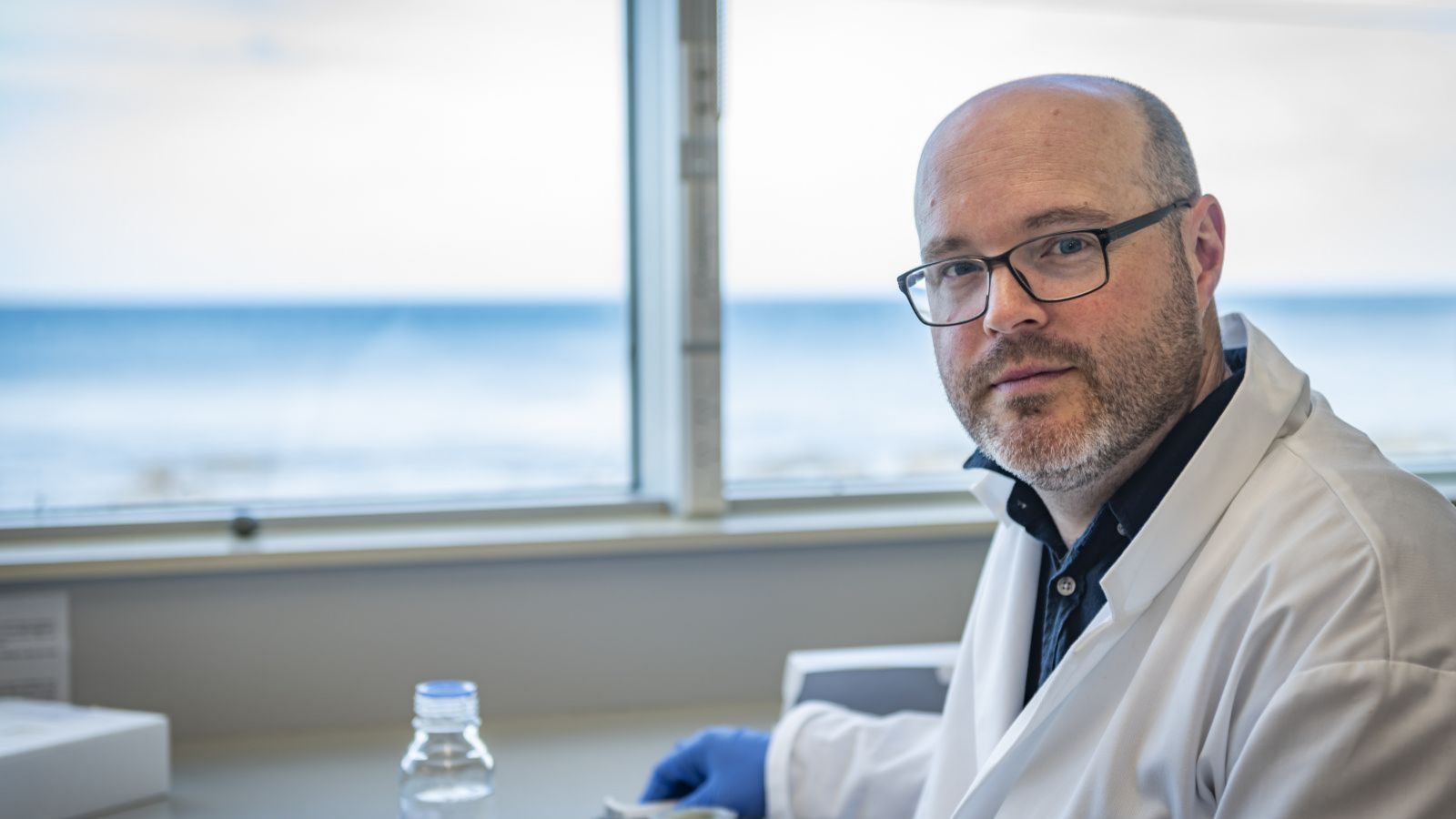 A man wearing glasses and a lab coat looks into the camera, with a background of the ocean.