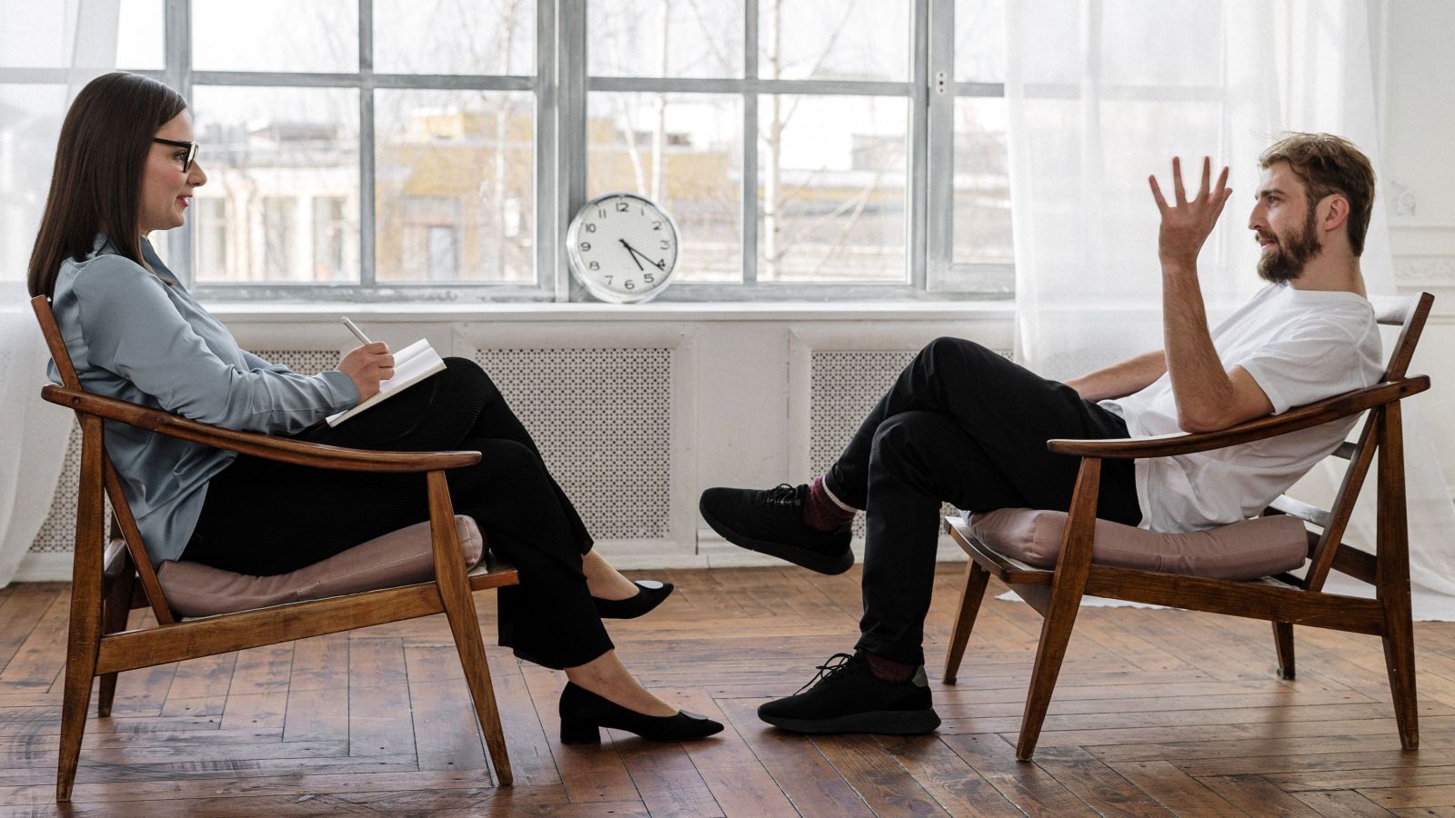 woman and man sitting in uncomfortable chairs with a clock in the background