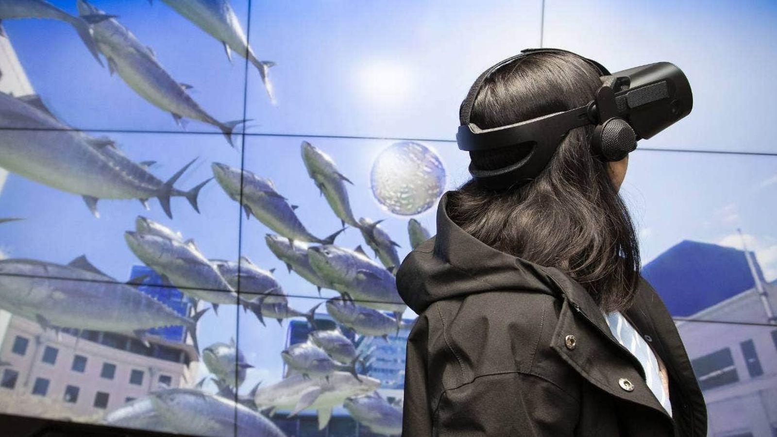 Woman wearing VR headset, screen behind her shows fish flying in sky