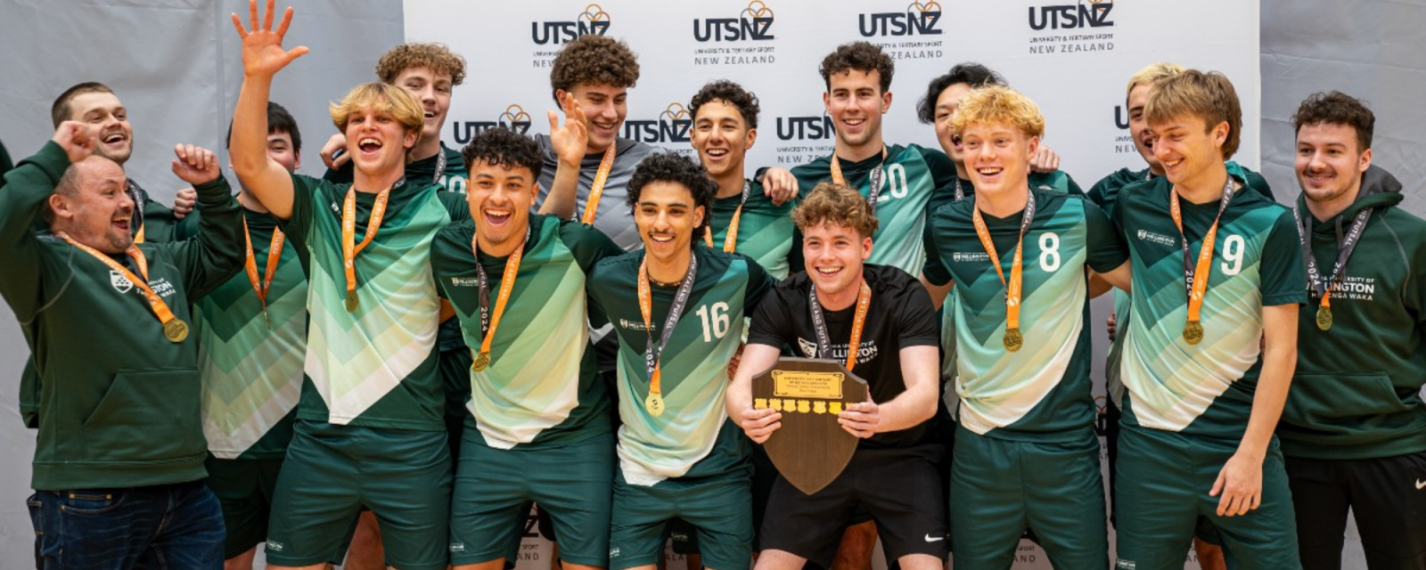 A group of students and staff pose together, holding a trophy at a sports tournament.