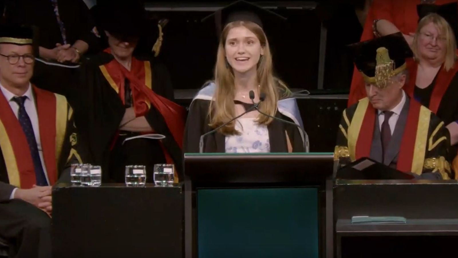 A smiling graduate wearing academic robes speaking at the ceremony.