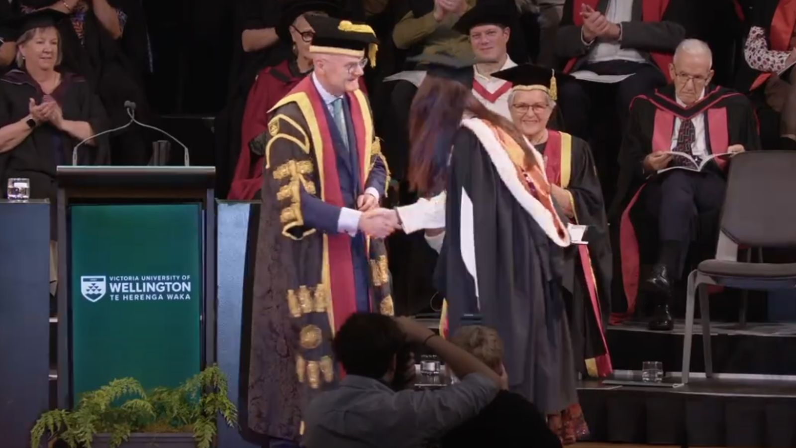 A graduate in regalia shakes hands with the Chancellor on stage during a graduation ceremony.