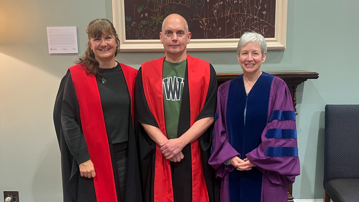 Three academics, one male, two female wearing academic dress in front of a pale green wall