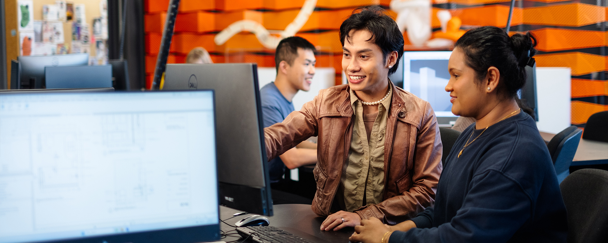 Two students in front of a computer screen, one is gesturing to the screen and advising the other student. There are other students on computers in the background.