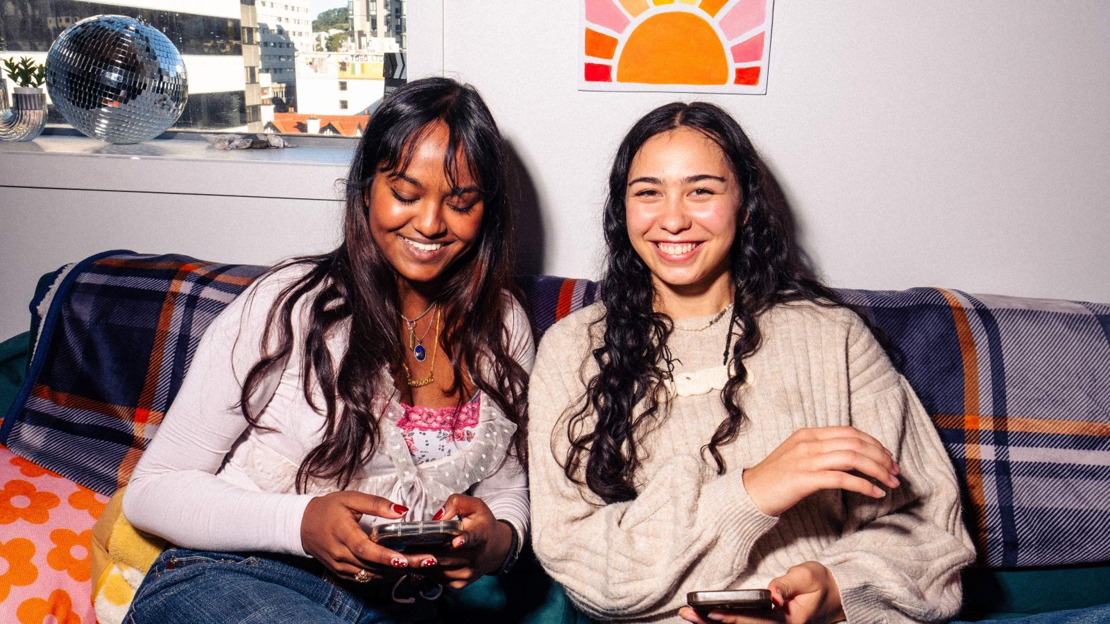 Two people sitting on a couch using smartphones in student accommodation. Behind them is a window showing a glimpse of Wellington CBD, there's a disco ball, a colourful blanket, and a sun-themed poster on the wall.