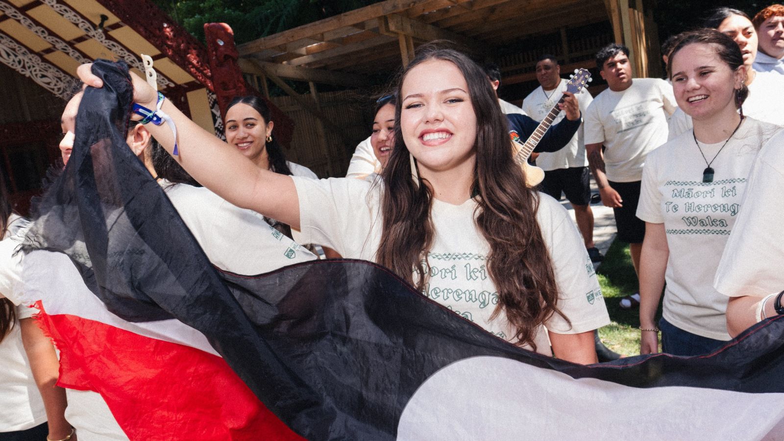 A group of students at marae, there is a person at the front of the group waving a flag and others behind them playing instruments