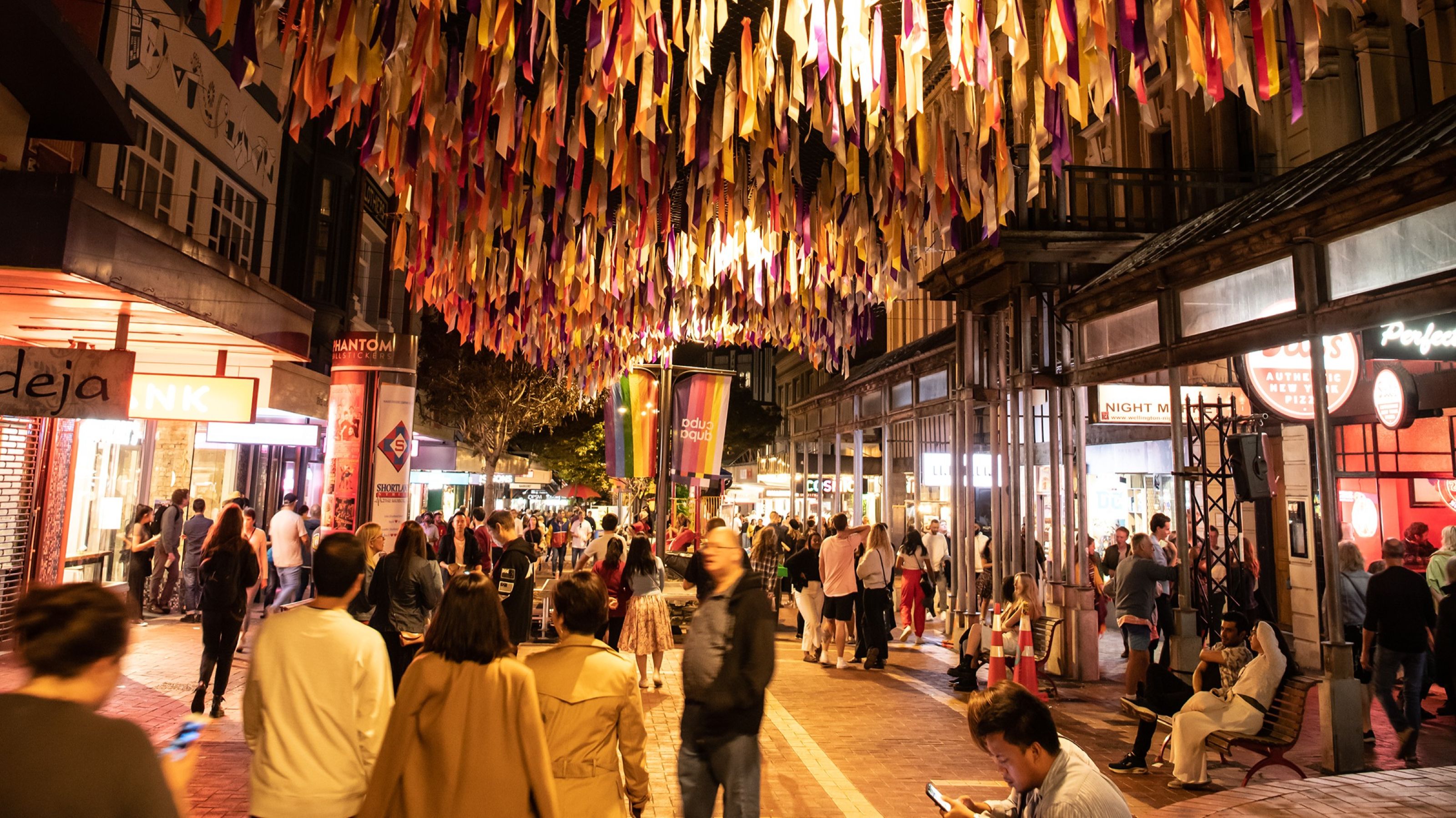 Busy pedestrian street at night, decorated with colourful hanging ribbons overhead, with crowds of people walking among lit shops and restaurants and rainbow banners visible along the street.
