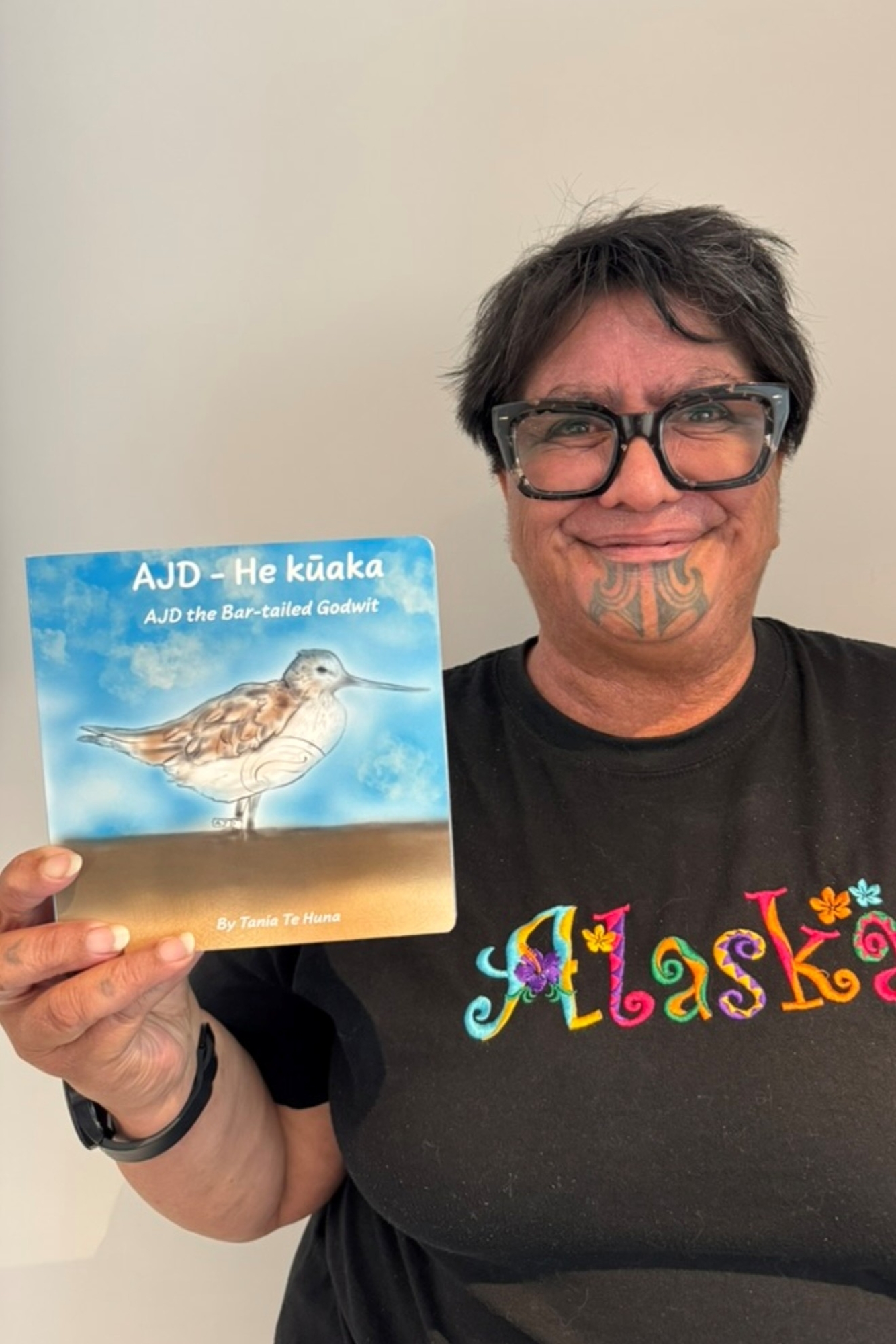 Tania Te Huna stands holding a children's book with an illustration of a bird on the front.