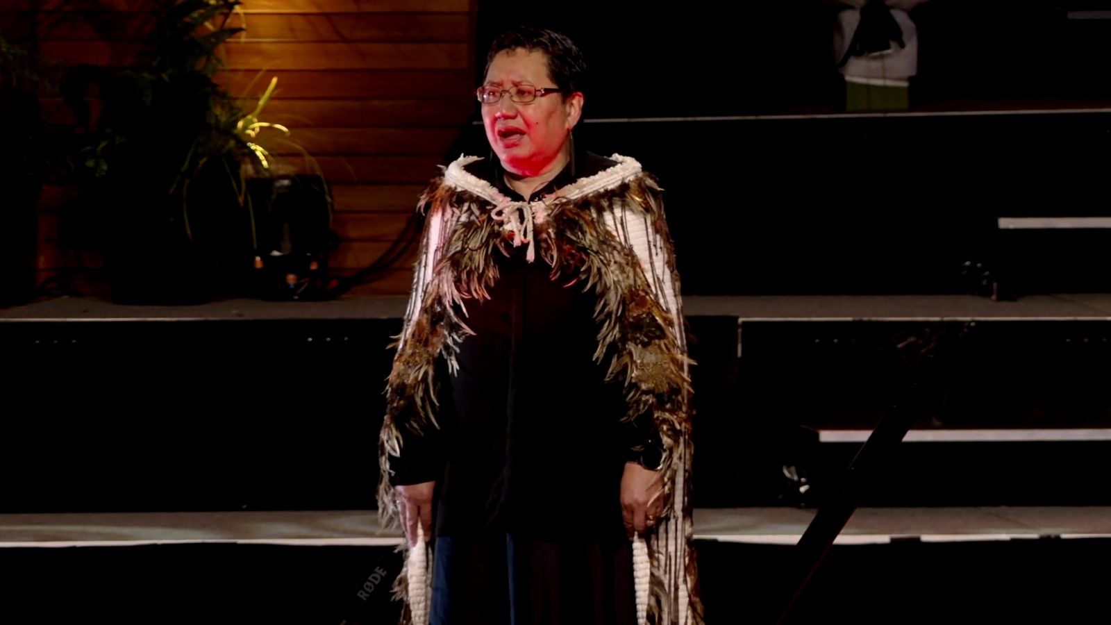 Person wearing a feathered cloak performing on stage during a graduation ceremony.