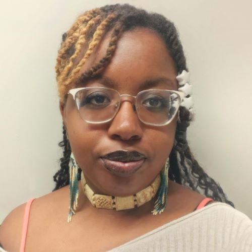 Person wearing glasses, a beige top, and patterned earrings, standing in front of a beige background.