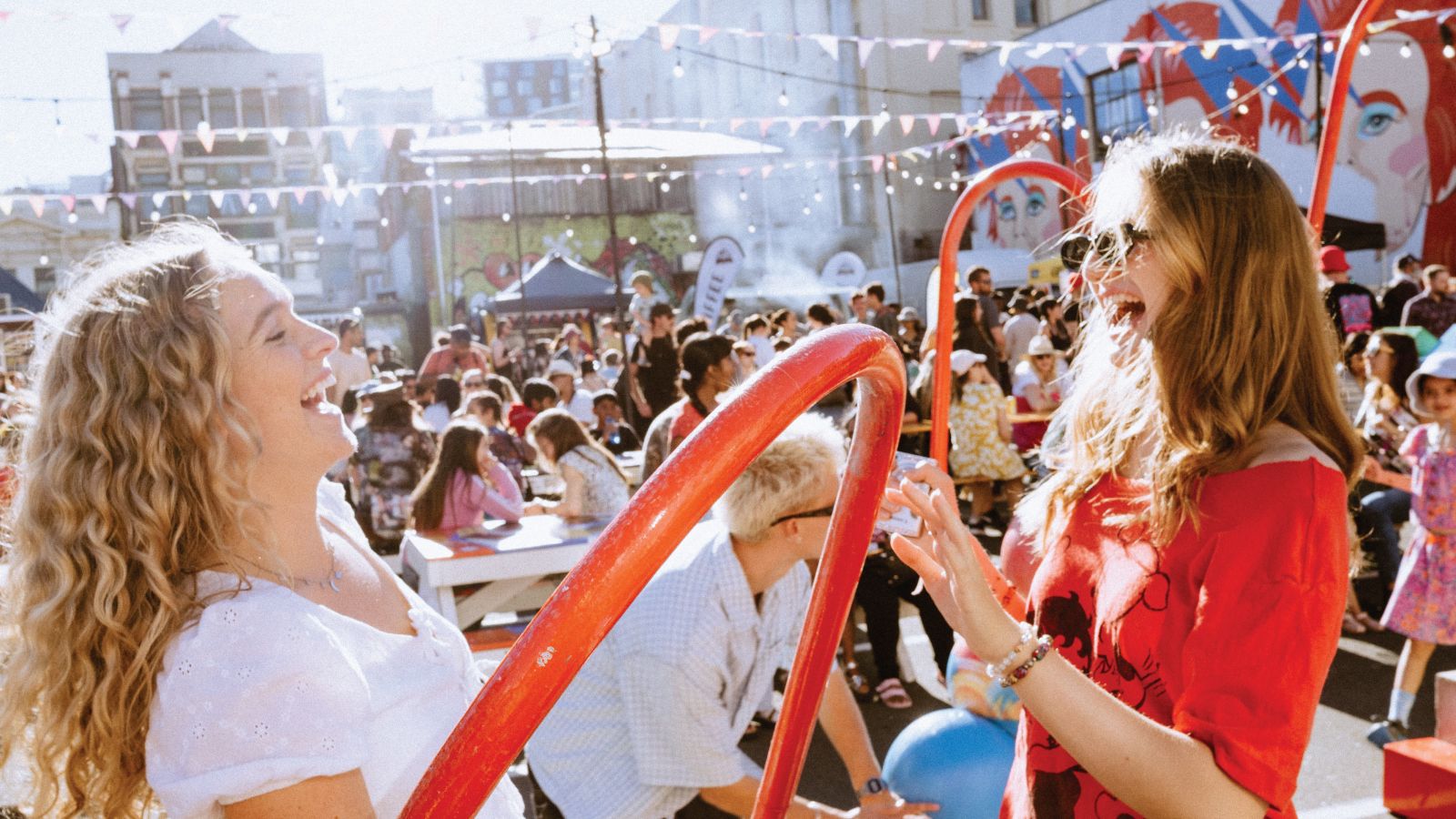 2 students at a street party, laughing and using a hula hoop, with large groups of people sitting on benches behind them