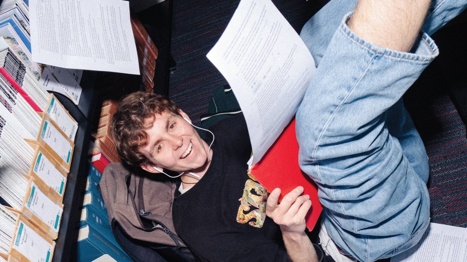 Male student sitting on the floor in the library, reading and smiling. Study papers are flying in the air.