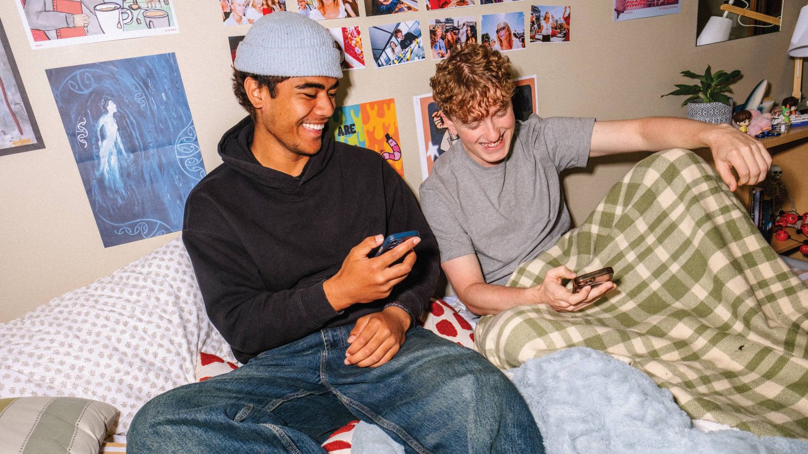 Two male students sitting on a bed in halls of residence looking at their phones and smiling,