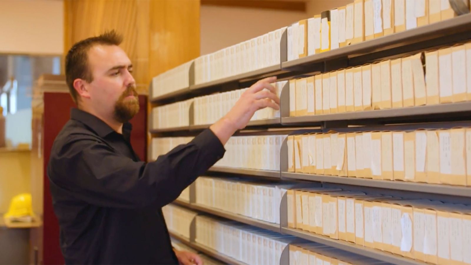 A man selects a small case from shelves of contianing the same sized cases in an archive.