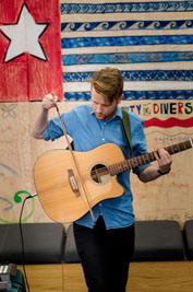 Blake stands in front of a wall decorated with drawings playing the guitar with a violin bow.