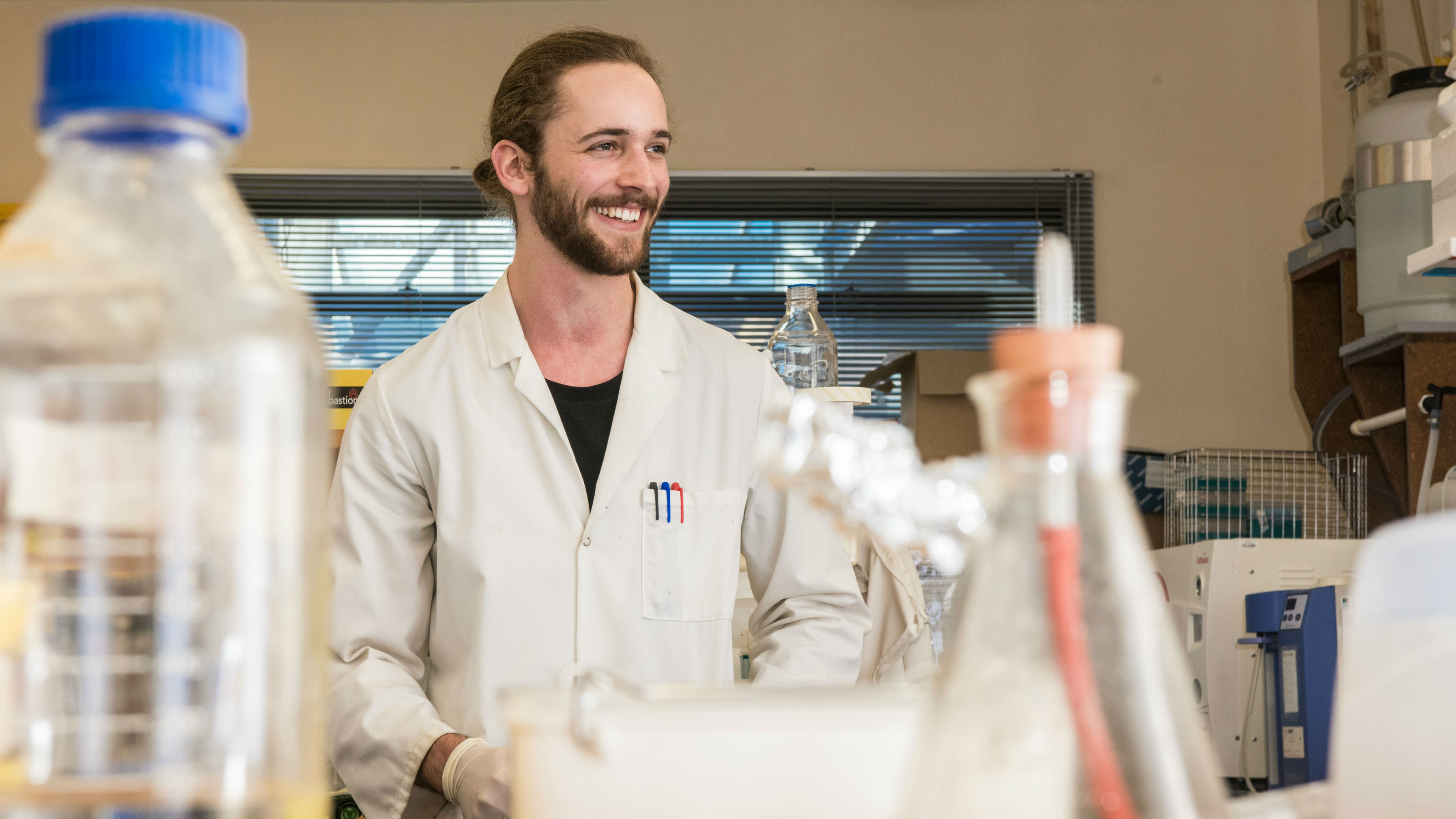 A student wearing a white lab coat stands smiling over science eqiupment in a laboratory.