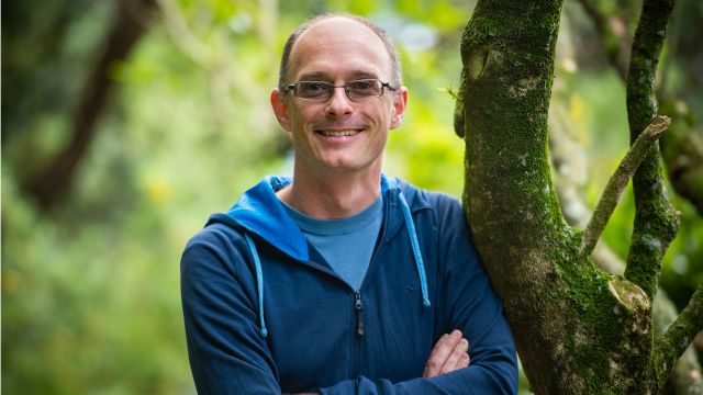 Stephen Marsland, wearing a blue hoodie, leans against a tree with green vegetation behind him. 
