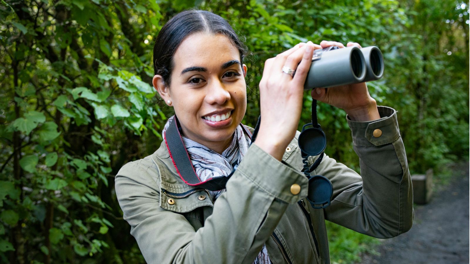 Gliselle Marin holds up binoculars with green trees and bush behind her. 