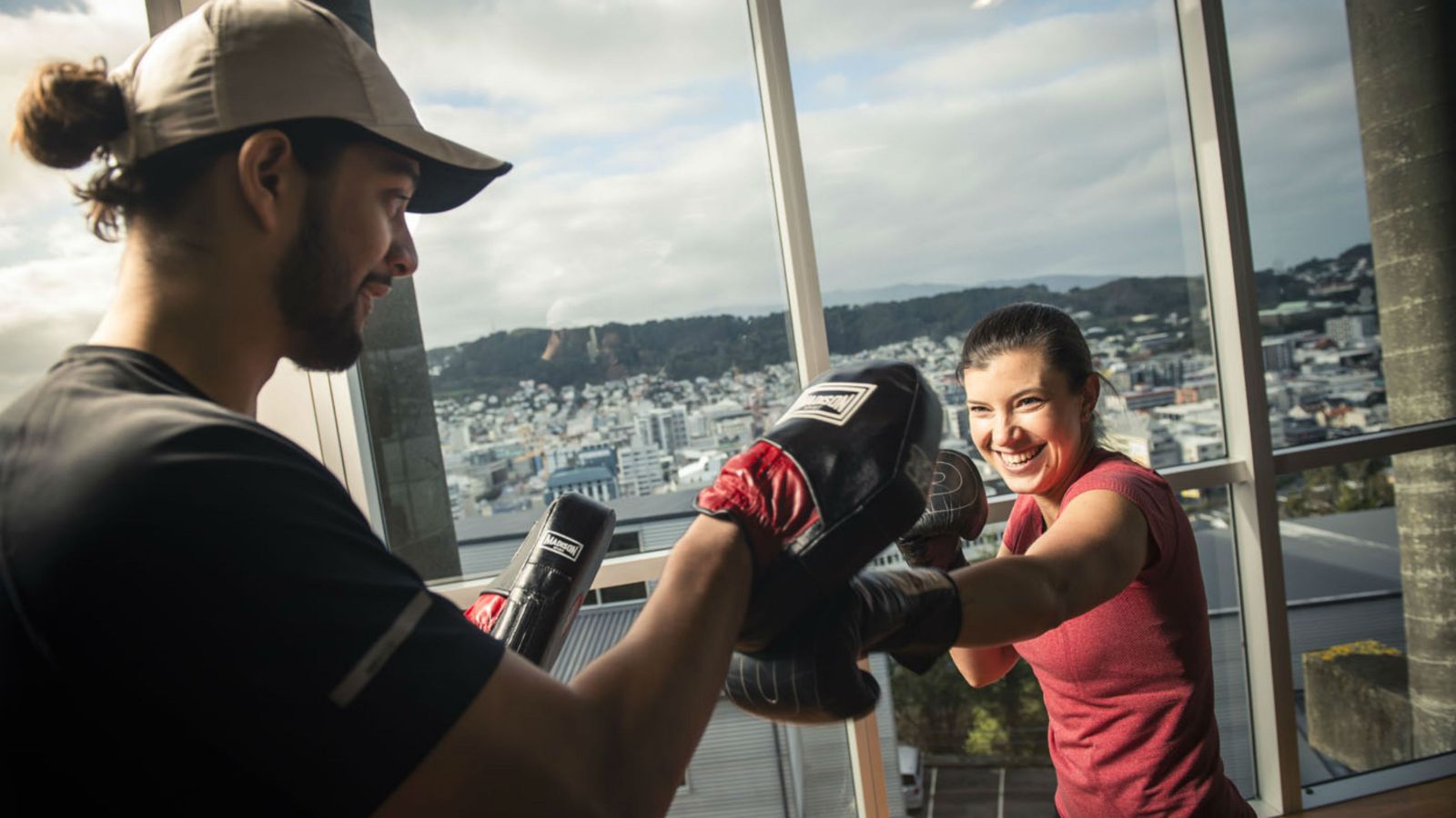 A female student with boxing gloves, punches practice targets held up by a male instructor.