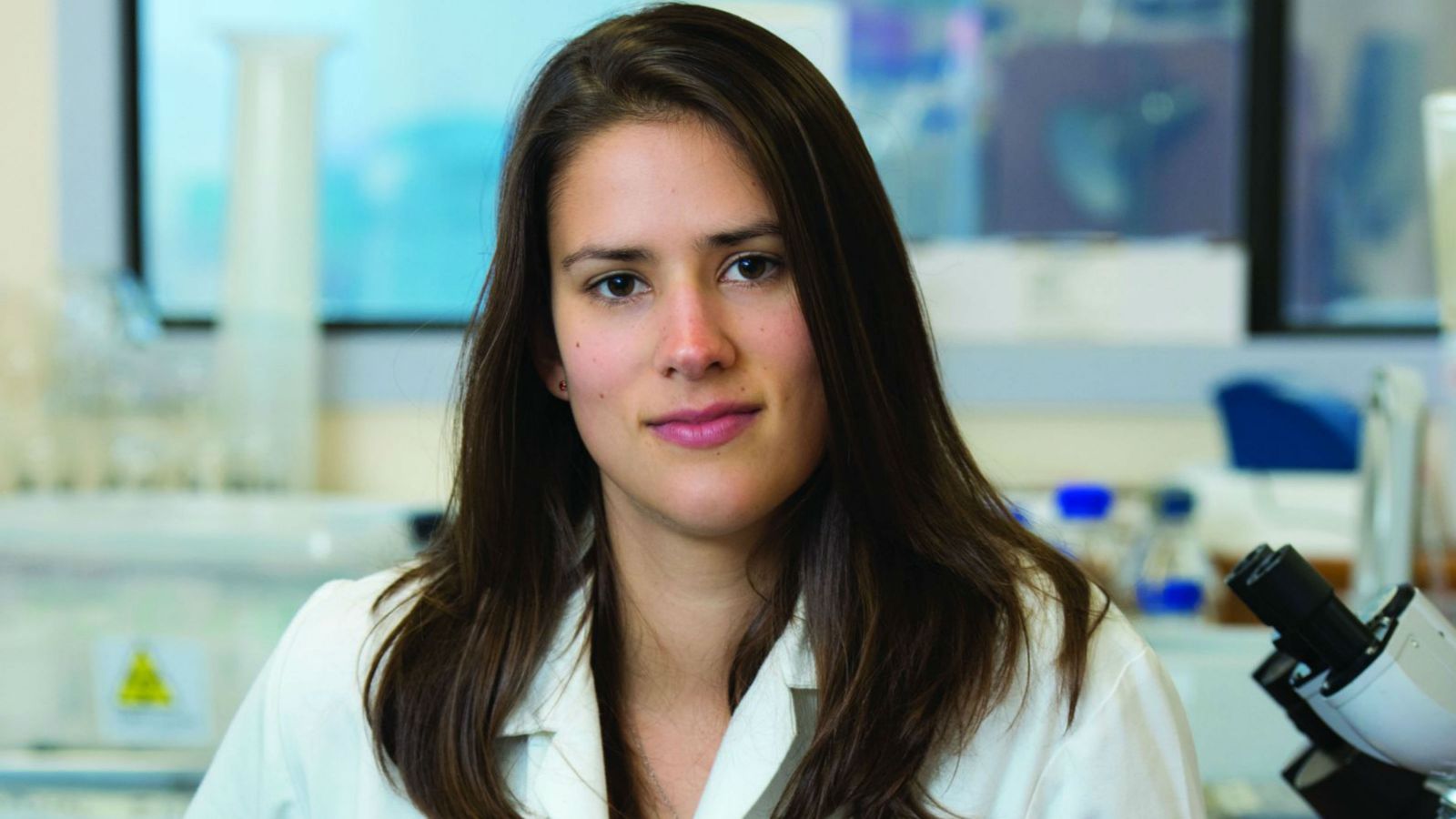 Jennifer, wearing a lab coat, stands next to a microscope and looks directly at the camera. There is blurred lab equipment in the background. 