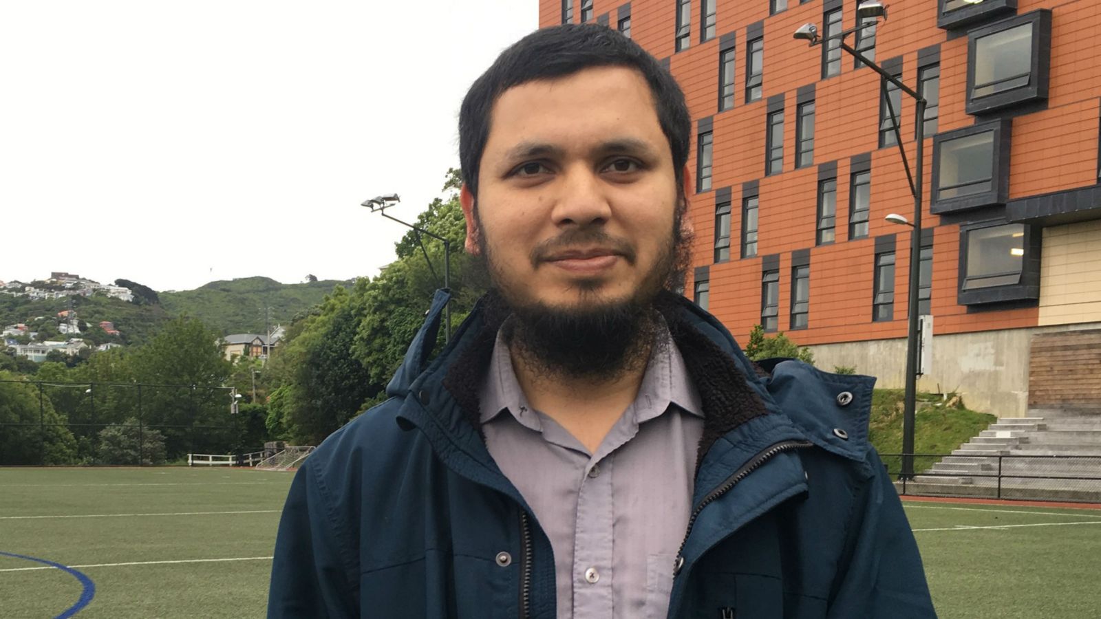 Arif stands in the middle of a sports field, with rolling hills and an orange building behind him. 