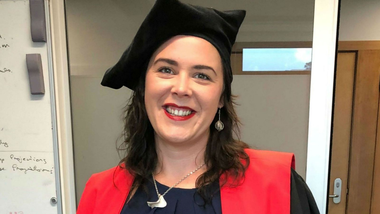 Georgia, wearing her graduation regalia, looks directly at the camera with a white board and door behind her. 