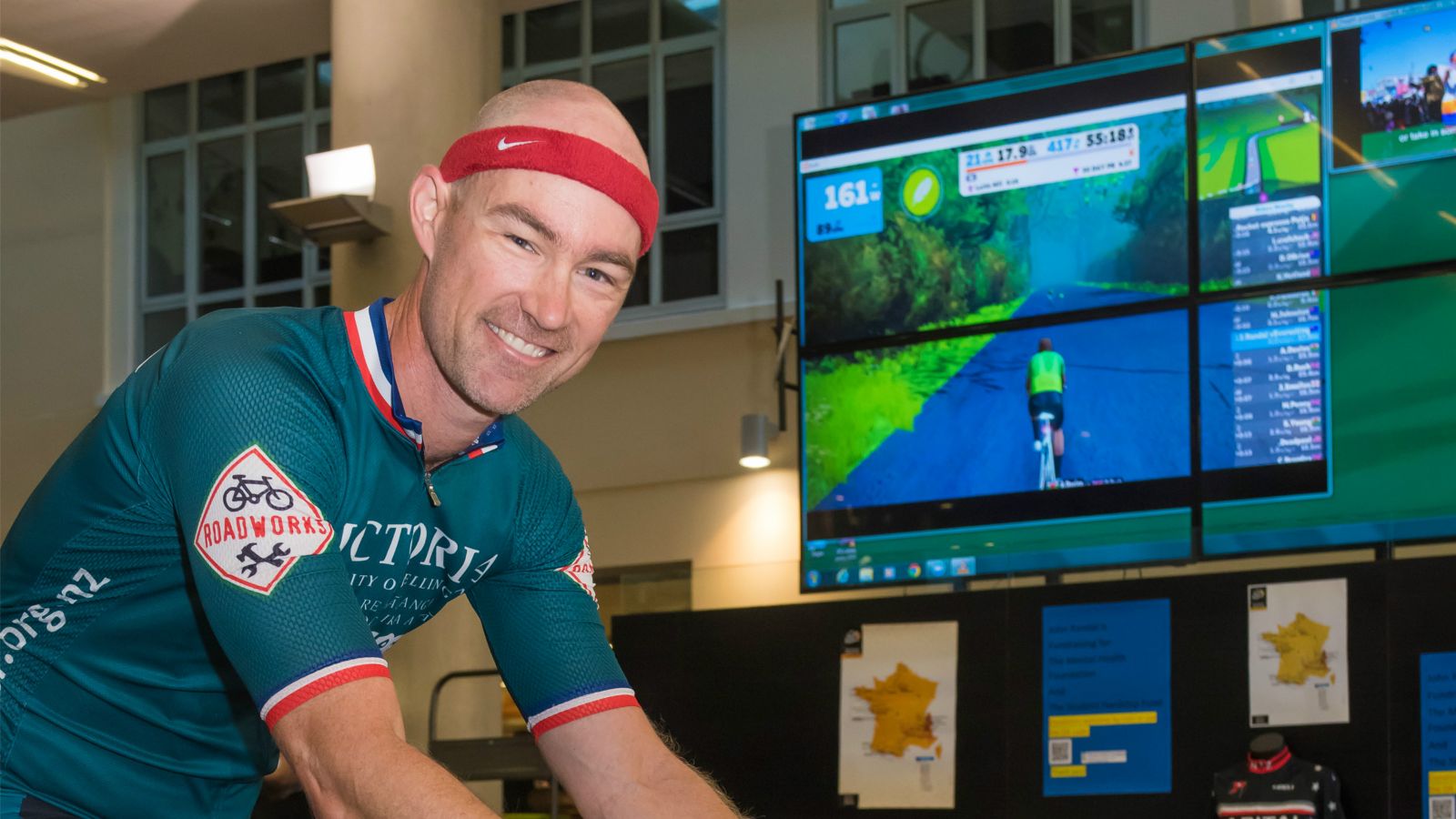 Dr John Randal rides a stationary bicycle with an image of a bicycle in an interactive simulation is presented on a large screeen.