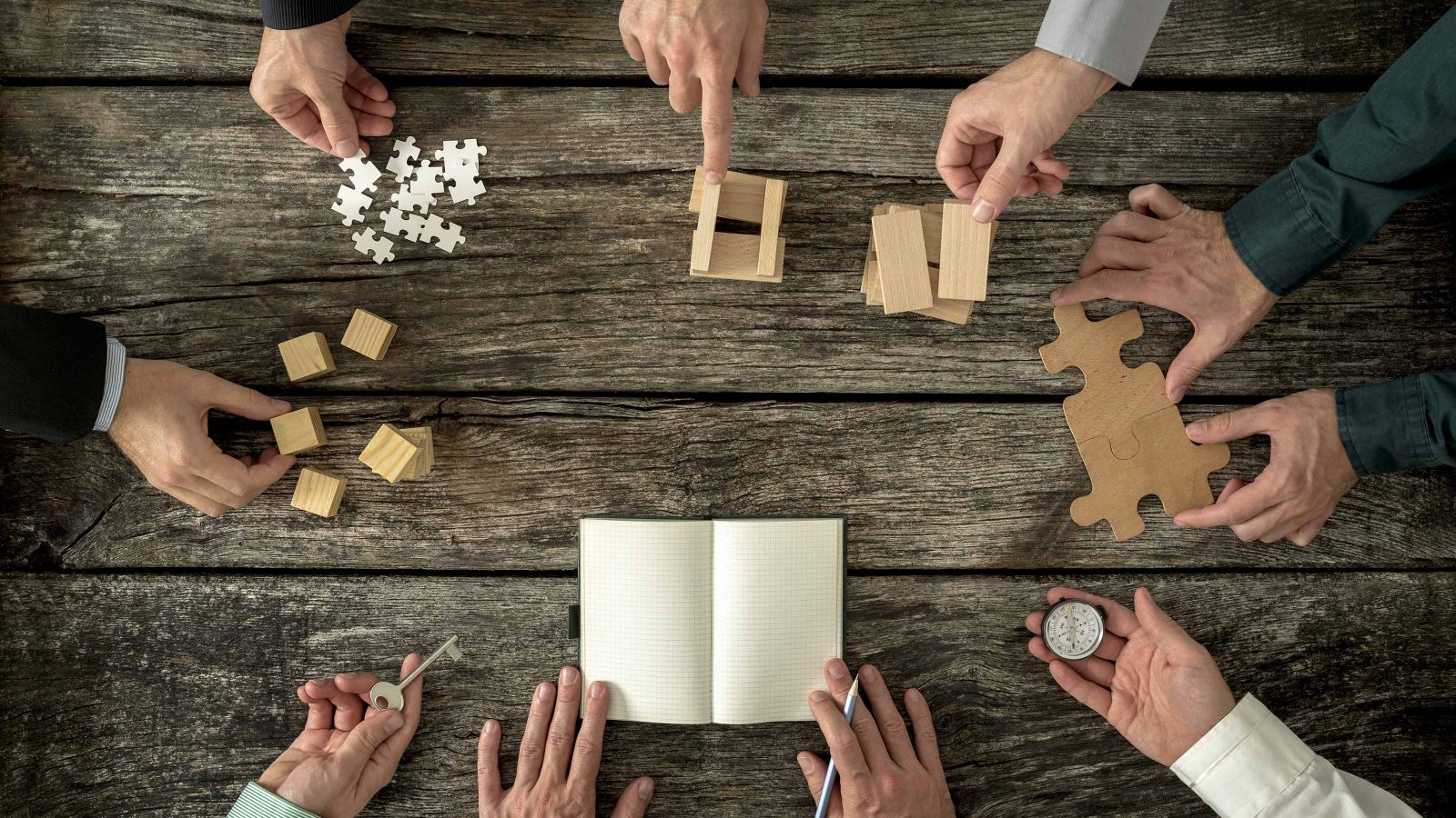 An image of hands around a wooden table holding various small objects, like a compass, small cubes, puzzle pieces, a key and a note book.