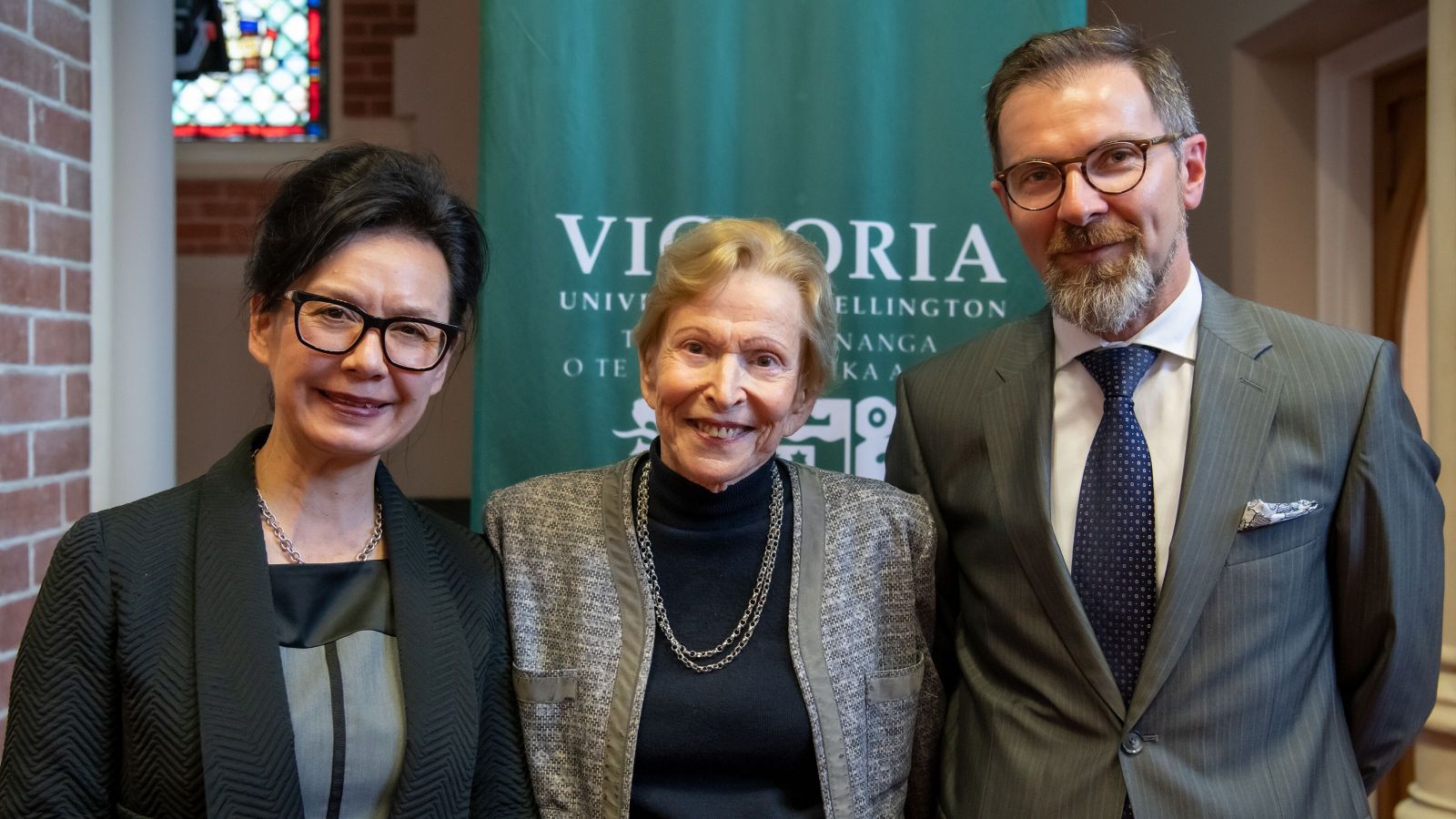 Two women and a man in front of a banner with victoria university on