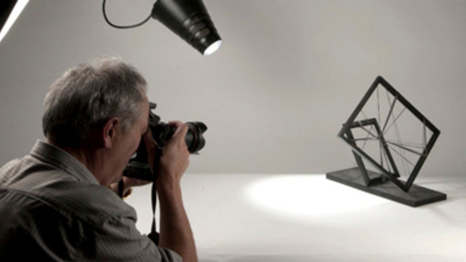 A grey-haired man photographs an abstract frame shape in a light grey studio with a single spotlight.