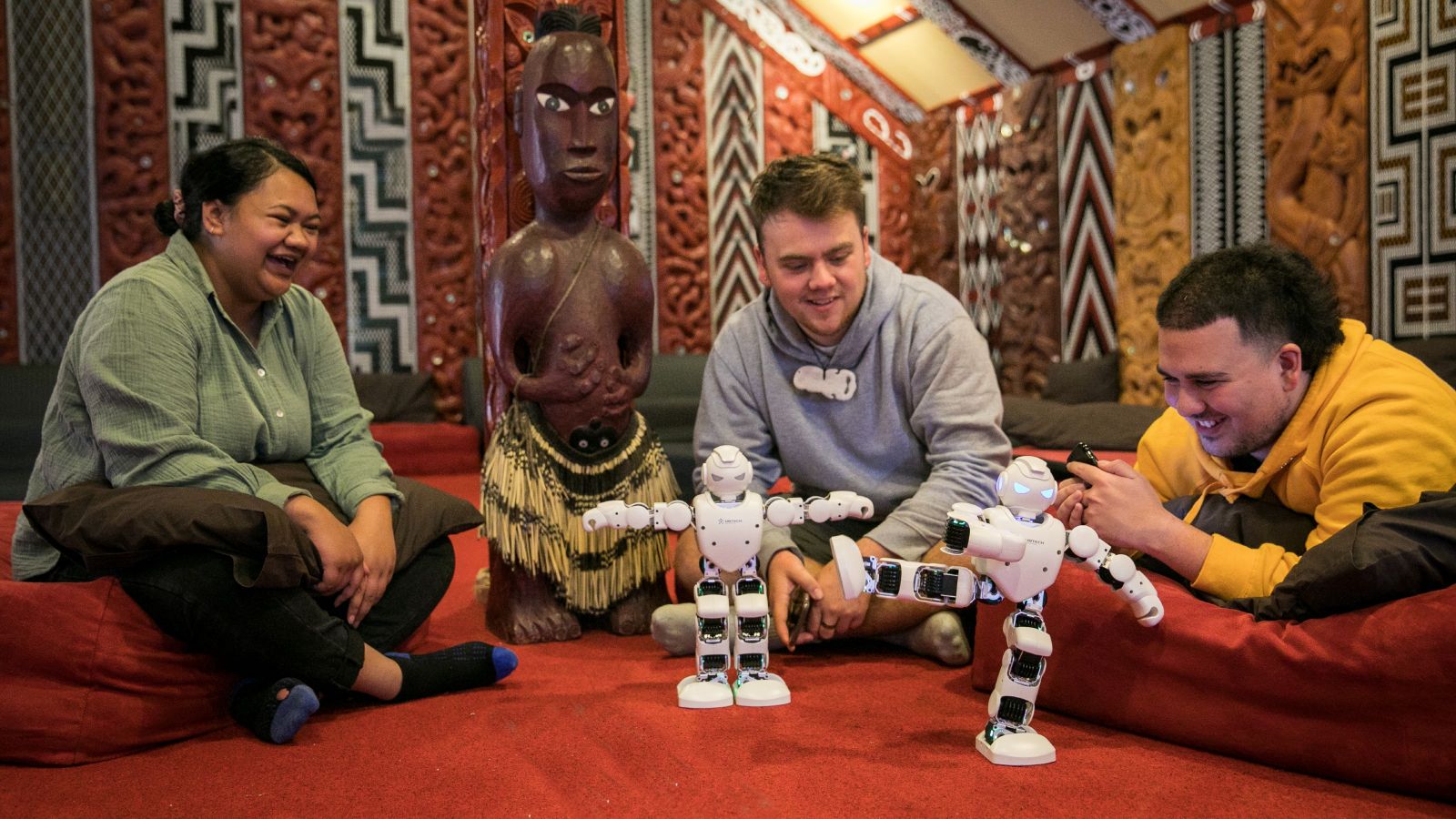 Three students sitting in Te Herenga Waka marae playing with two robots.