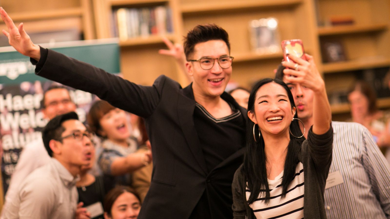 Man and a woman smiling and taking a selfie, with crowd of people posing for the photo in the background.