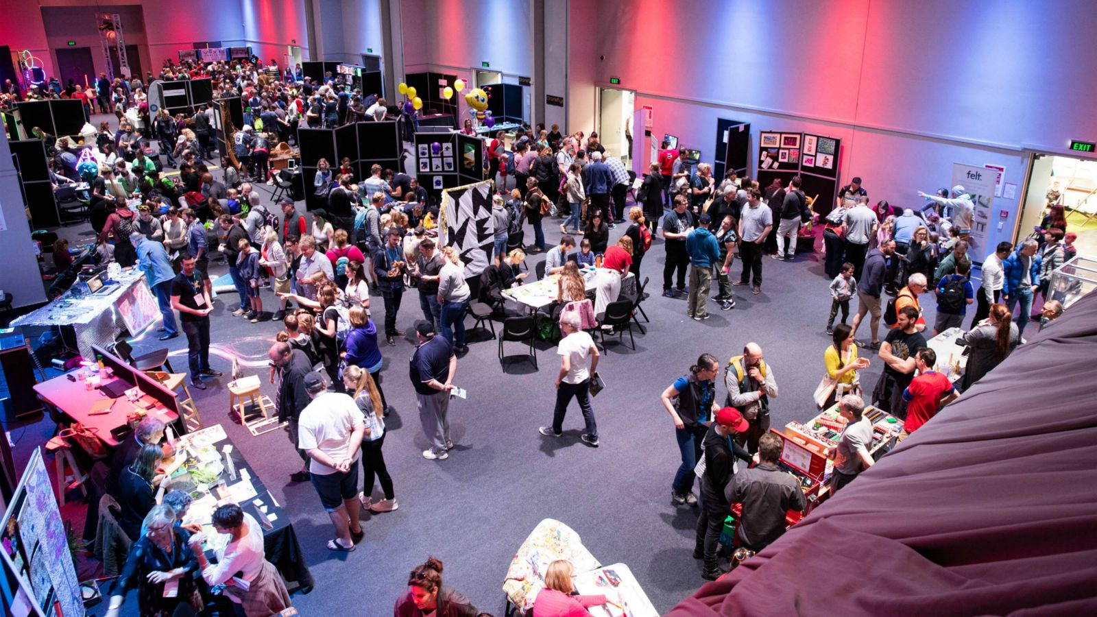 Maker Faire Wellington in full swing. Photo credit: Justine Hall – Large room lit in red and blue with people gathered around exhibition stands.