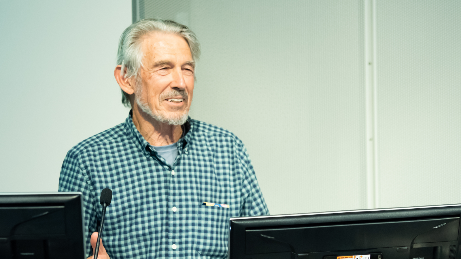 man with white hair and blue check top behind two screens giving a lecture