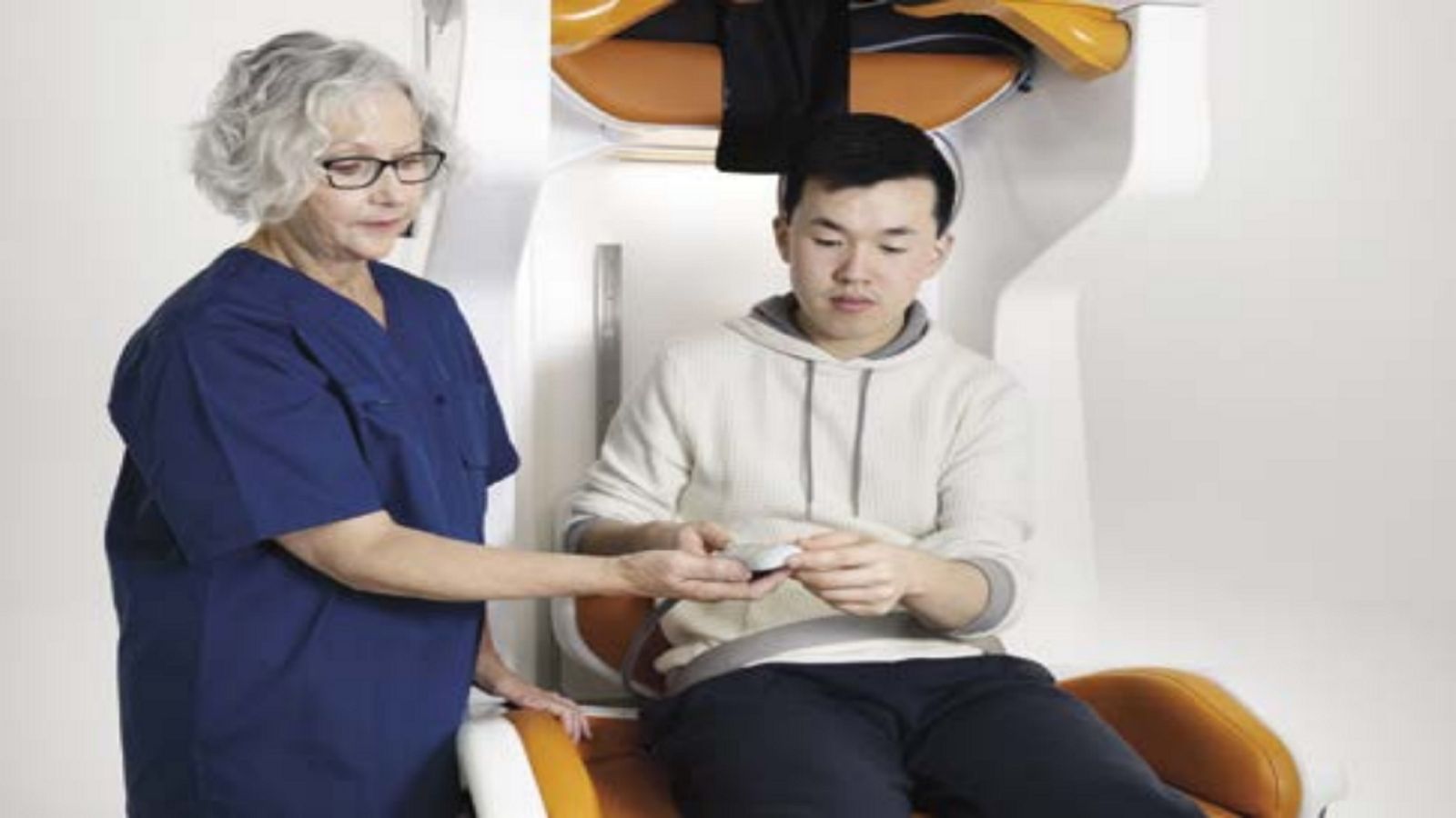 Man sitting in a portable MRI machine with a female medical professional in blue scrubs standing beside him.