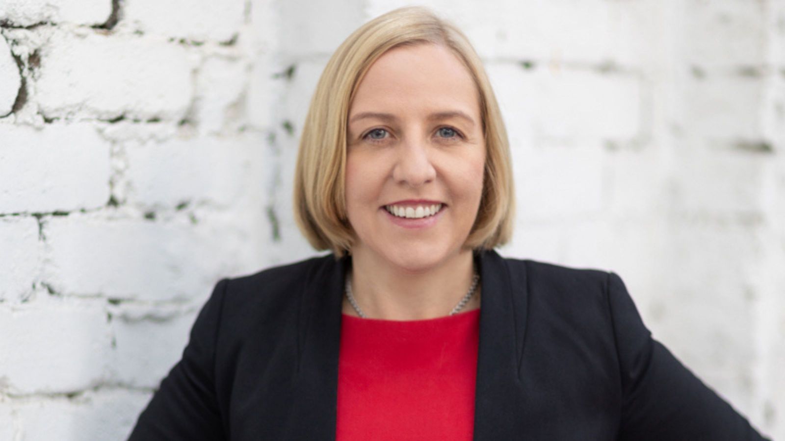 Woman with bobbed blond hair with red top and black formal jacket. 