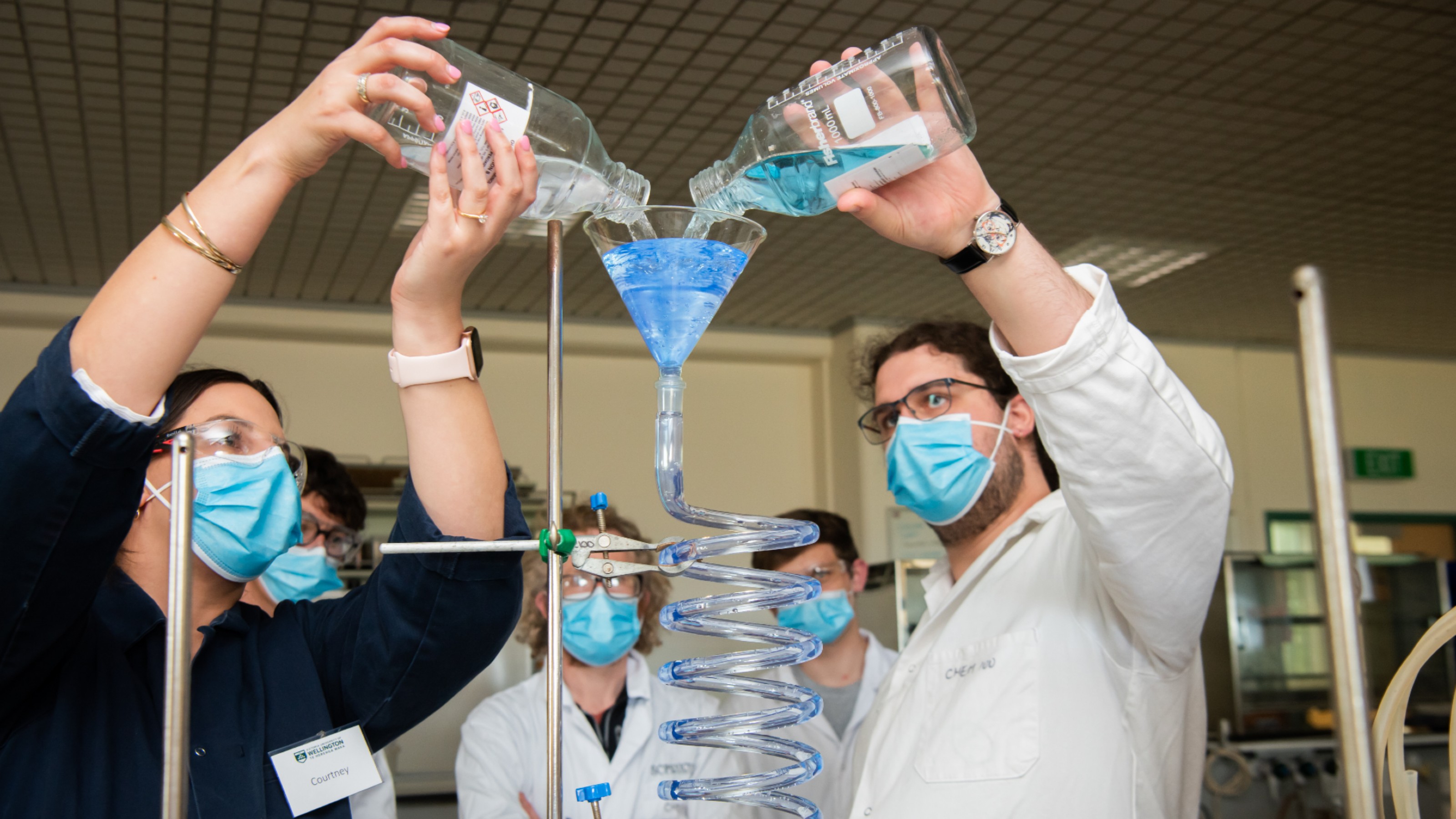 Two chemists in a lab pour blue liquid into a glass funnel