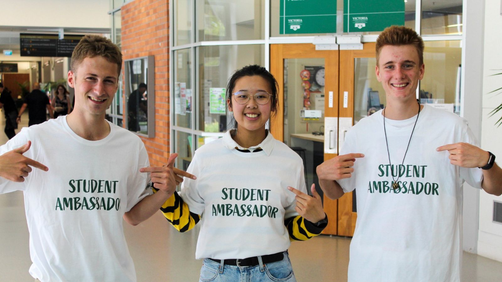 Three smiling students wearing T-shirts saying 'student ambassador' standing outside a faculty office.