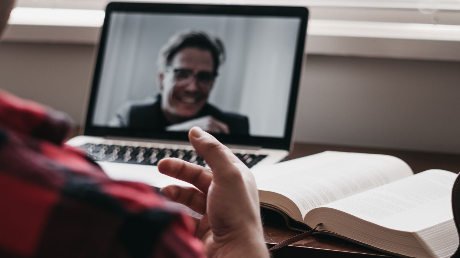 View from behind a student's shoulder as he gesticulates on a Zoom call on his laptop, next to a book.