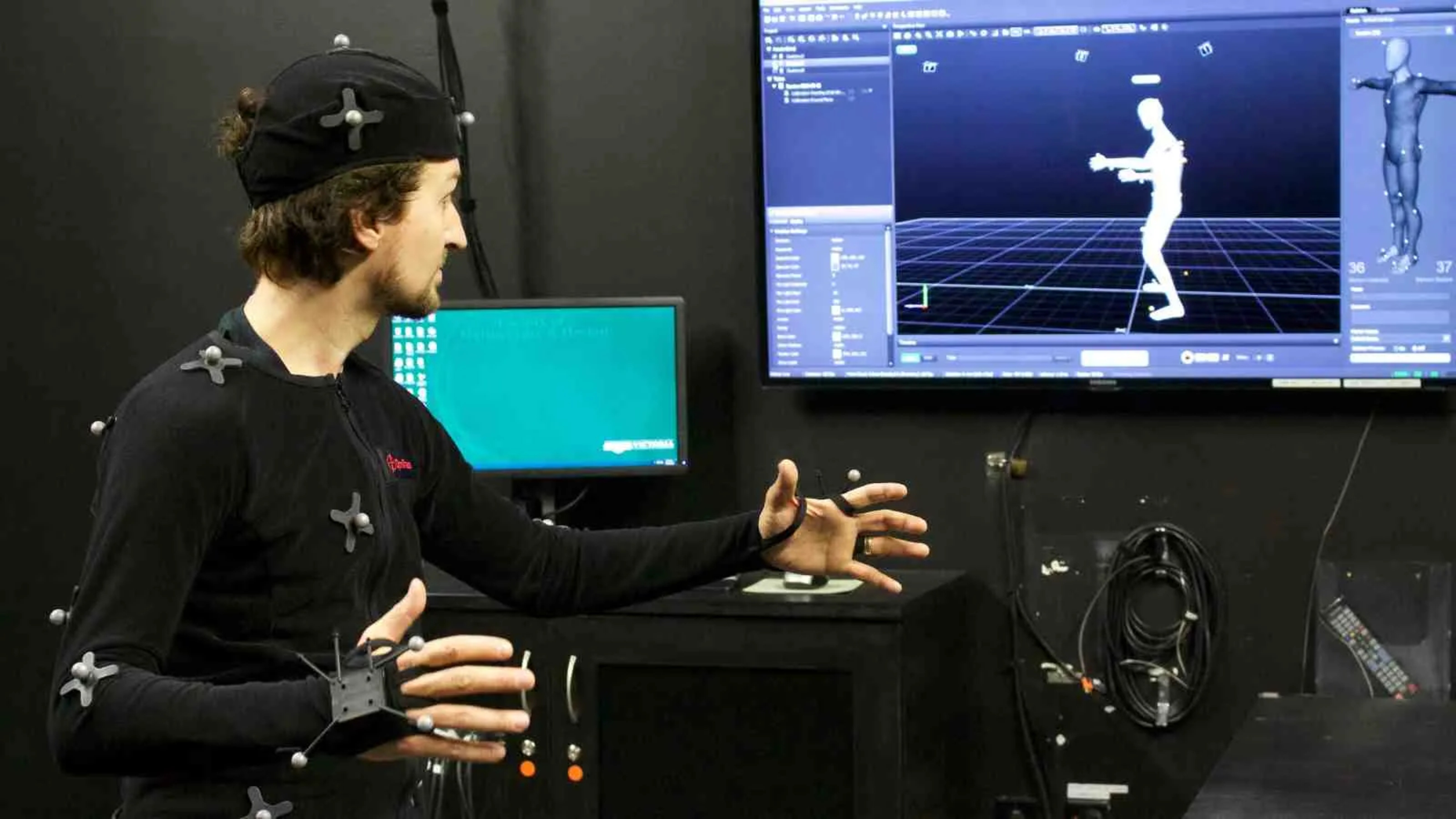 A man dressed in a black outfit covered in sensors looks at a monitor showing a computerised rendering of his body in a virtual space